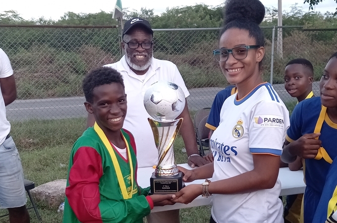 Deserine Harripaul (right), member of the YCDF organizing committee, presents the winner’s trophy to Old Harbour Primary football captain Roshane Reid. In background is Carl Cargill, YCDF founder. (OH News Photo)